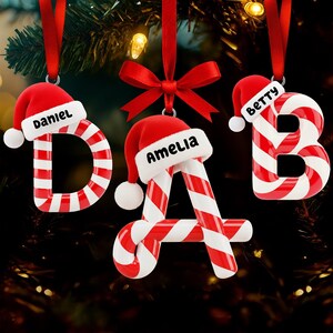 May include: Christmas ornaments shaped like the letters D, A, and B, each with a red and white candy cane design. Each letter is topped with a red Santa hat with a white pom-pom and a name printed on the hat. The ornaments hang from red ribbons.
