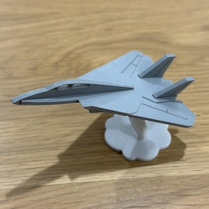 May include: A light grey model fighter jet with a sleek, angular design and a pointed nose. The aircraft features dual vertical stabilisers and is mounted on a white, cloud-shaped base that provides a stable display for the miniature vehicle.