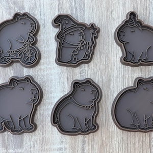 Könnte beinhalten: Sechs dunkelgraue Ausstecher in Form eines Capybaras. Die Designs zeigen ein Capybara auf einem Fahrrad, als Zauberer verkleidet, mit Partyhut und in anderen Posen. Ideal für thematische Kekse.