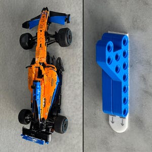 May include: An overhead shot of a detailed orange and blue race car model, alongside a blue plastic mounting bracket. The race car has black wheels and intricate detailing. The bracket has multiple holes and a white adhesive strip.