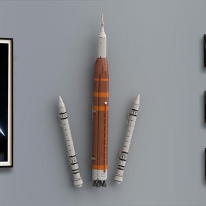 May include: A vertical wall-mounted model of a space launch vehicle featuring a central orange and white cylindrical rocket body flanked by two thinner white booster rockets, all displaying detailed mechanical segments and engine nozzles.
