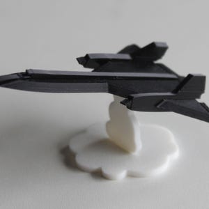 Puede incluir: Un modelo de avión de combate furtivo de plástico negro sobre una base blanca en forma de nube.