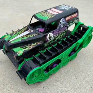 Könnte beinhalten: Ein schwarzes und grünes Grave Digger Monster Truck Spielzeug mit einem Totenkopf-Motiv auf dem Dach. Der Truck hat grüne Flammenmuster und ist auf grünen Panzerketten montiert. Das Spielzeug ist aus Kunststoff.