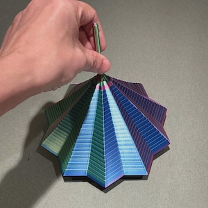 May include: A multi-colored, geometric paper ornament with a green stem. The ornament features a starburst design with shades of blue, green, and purple. The paper is folded into a series of triangular shapes, creating a layered effect. The stem is held by a hand.