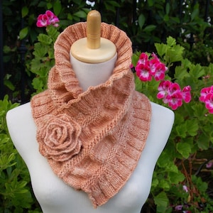 May include: A peach-colored knitted cowl with a large crocheted flower accent. The cowl is displayed on a white mannequin against a backdrop of green foliage and pink flowers.