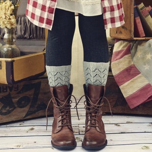 Op de afbeelding: Een paar bruine leren laarzen met veters en een grijze gebreide manchet. De laarzen worden gedragen door iemand die een rood-wit geruit overhemd en een witte rok draagt.