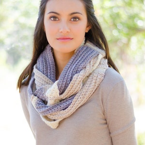 May include: A woman wearing a long-sleeved, light brown jumper with a crocheted scarf in shades of beige, white, and purple. The scarf is wrapped around her neck and hangs down her chest.