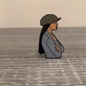 May include: A pin featuring a side profile of a person wearing a gray blazer, a dark green cap, and large hoop earrings. The person has long black hair and crossed arms. The pin is set against a gray wooden surface.
