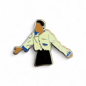 May include: Enamel pin depicting a stylized figure in a light yellow jacket with blue details and a black skirt. The pin has a gold outline and shows a person with arms outstretched.