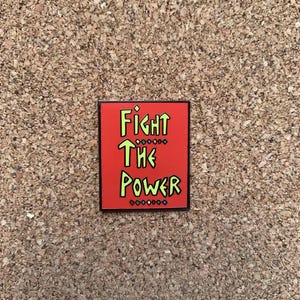 May include: A rectangular pin with the words "FIGHT THE POWER" in neon green lettering on a red background. The pin has a black border and is set against a corkboard.