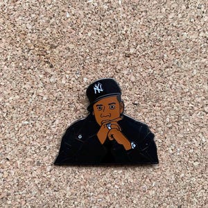 May include: A black enamel pin featuring a stylized illustration of a man wearing a black cap with a white NY logo. The man is wearing a black jacket and has his hand near his mouth.