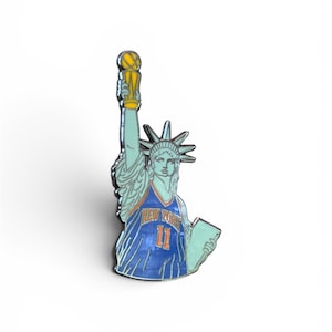 Könnte beinhalten: Eine Metall-Emaille-Anstecknadel, die die Freiheitsstatue in einem blau-orangen Basketballtrikot mit dem Text New York und der Nummer 11 zeigt. Die Figur hält einen gelben Basketball-Pokal in der einen und eine Tafel in der anderen Hand.