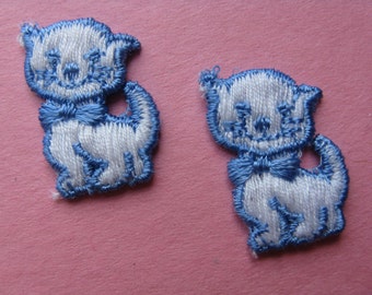 May include: Two white embroidered cat appliques with blue details. The cats are standing with their tails curled behind them. They have blue bows around their necks.