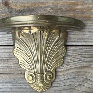 May include: A wall-mounted shelf crafted from polished brass, featuring a semi-circular top surface and a decorative support bracket shaped like a large, vertically oriented seashell with intricate ribbed detailing and scrolled accents at the base.