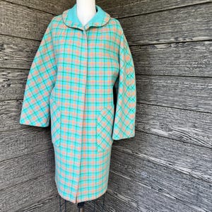 May include: A long-sleeved coat featuring a vibrant turquoise and peach plaid pattern. The garment includes a rounded collar, a button-front closure, and two large patch pockets on the front. The coat has a straight silhouette and reaches below the hips.
