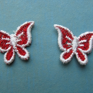 May include: Two red and white embroidered butterfly appliques. The butterflies have a white outline and red stripes.