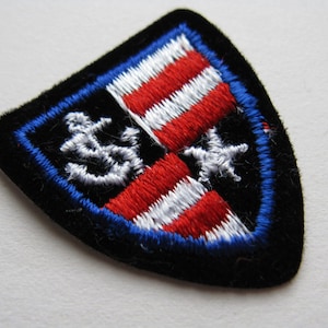 May include: A black felt patch with a red, white, and blue embroidered American flag design. The flag is centered on a shield shape with a white anchor and a white star on either side of the flag.