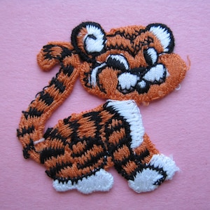 May include: Orange and black embroidered tiger applique with white accents. The tiger is facing right and has a cartoonish expression.