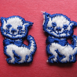 May include: Two white felt cat appliques with blue embroidered details, including a bow tie. The cats are sitting with their tails curled around their bodies.