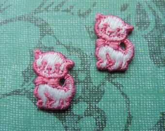 May include: Two white felt cat appliques with pink embroidered details. The cats are standing upright with their tails curled around their bodies.