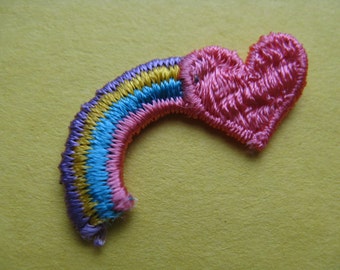 May include: A rainbow-colored embroidered patch shaped like a heart. The heart is pink and the rainbow is made of purple, yellow, blue, and pink thread.