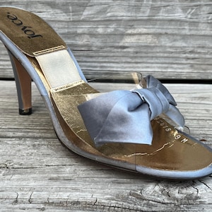 May include: A vintage high-heeled sandal with a clear strap and a bow detail. The shoe features a gold insole, a light blue exterior, and a stiletto heel. The brand name "Joyce" is visible on the insole.