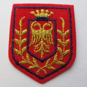 May include: A red embroidered patch with a shield shape. The patch features a gold emblem of a double-headed eagle, a crown, and floral elements, all outlined in blue thread. The patch is likely a decorative emblem or insignia.