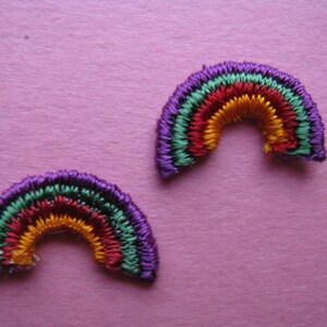 May include: Two embroidered rainbow patches. Each patch features a semi-circular design with bands of purple, green, red, orange, and yellow thread. The patches are set against a pink background.
