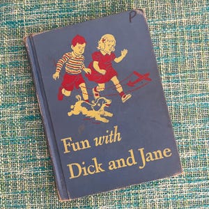 May include: A vintage hardcover book titled Fun with Dick and Jane with a dark blue cover. The front features a graphic illustration of a boy and girl running with a small dog and a red toy airplane. The letter P is written in the top corner.