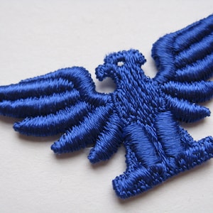 May include: A blue embroidered eagle applique patch. The eagle has its wings spread out and is facing to the right. The patch is made of a soft, textured fabric.