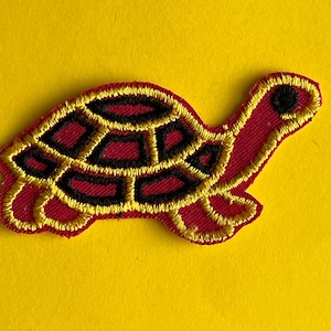 May include: Embroidered turtle patch in red, black, and gold. The turtle's shell is black with gold outlines, and the body is red with gold edging. The patch is on a yellow background.