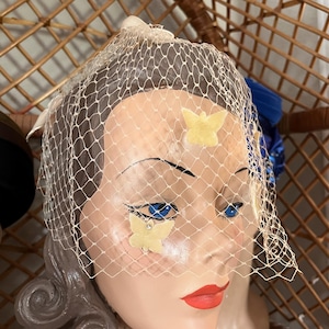 May include: A mannequin head with a vintage-style veil. The veil is a fine, cream-colored netting, adorned with small, pale yellow butterfly appliqués. The mannequin has blue eyes and red lipstick. The background includes a wicker structure.