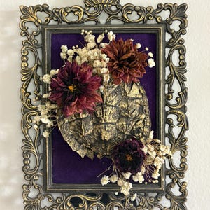 May include: A decorative wall hanging featuring a gold-painted turtle shell mounted on a deep purple velvet background within an ornate, antique-style gold metal frame, accented with dried dark red dahlias and small white baby's breath flowers.