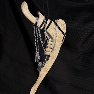 May include: A beige animal jawbone pendant suspended by a black cord, featuring multiple silver-toned metal chains draped across the bone. Two clear, raw crystal points are attached to the ends of the silver chains, hanging vertically.