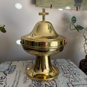 May include: A gold-colored, ornate chalice with a cross on top. The chalice has a rounded top and a flared base. The chalice is sitting on a surface with a map design.