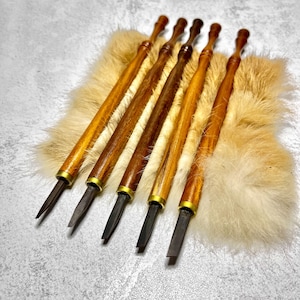 Może przedstawiać: Sześć drewnianych narzędzi do rzeźbienia z mosiężnymi tulejami i ostrymi ostrzami. Narzędzia są ułożone na powierzchni z białego i brązowego futra.