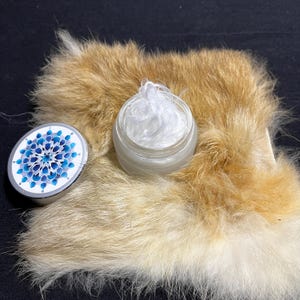 May include: A small white jar with a silver lid containing a white cream. The jar is sitting on a light brown fur surface.