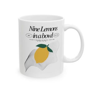 Może przedstawiać: Biały kubek ceramiczny z czarno-białym nadrukiem cytryny i tekstem "Nine Lemons in a bowl" z tekstem "SHOC" i "EST 2014" mniejszą czcionką.