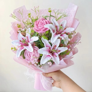 May include: A bouquet of crocheted flowers, featuring white lilies with pink accents, pink roses, and delicate greenery. The flowers are wrapped in pink paper and tied with a white ribbon. A handmade floral arrangement.