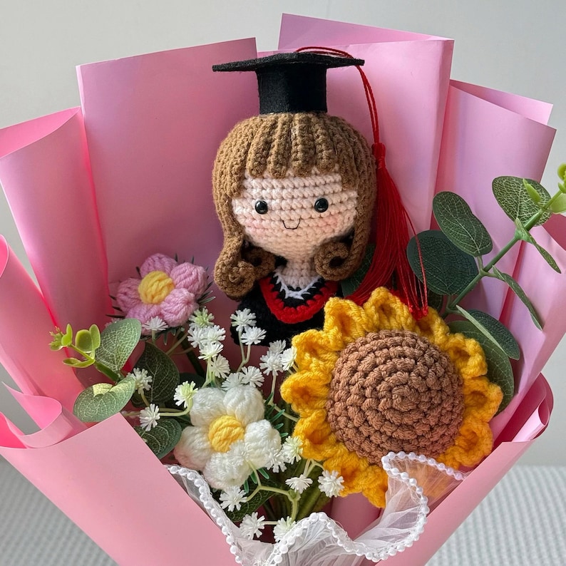 May include: A handmade crochet graduation bouquet featuring a doll wearing a black cap and gown. The bouquet includes crochet flowers in pink, yellow, and white, wrapped in pink paper. Greenery adds a natural touch to this unique gift.