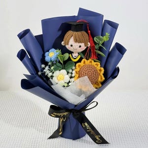 May include: A graduation-themed bouquet featuring a crocheted graduate doll wearing a black cap and gown, surrounded by crocheted flowers and greenery. The bouquet is wrapped in dark blue paper and tied with a black ribbon that says "Congratulations".