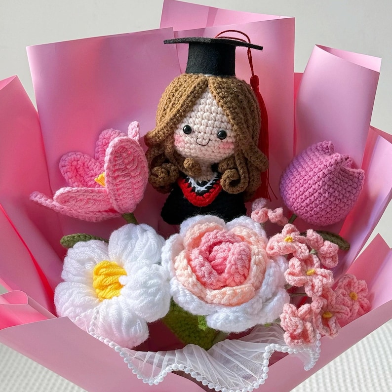 May include: A handmade crochet bouquet featuring a graduation doll wearing a black cap and gown. The bouquet includes pink, white, and yellow crochet flowers, wrapped in pink paper. A unique and thoughtful gift for a graduate.