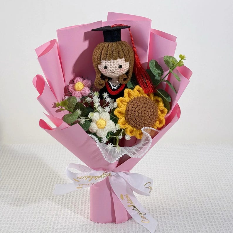 May include: A handmade graduation bouquet featuring a crocheted doll in a graduation cap, surrounded by crocheted flowers and greenery. The bouquet is wrapped in pink paper and tied with a white ribbon that says "Congratulations".