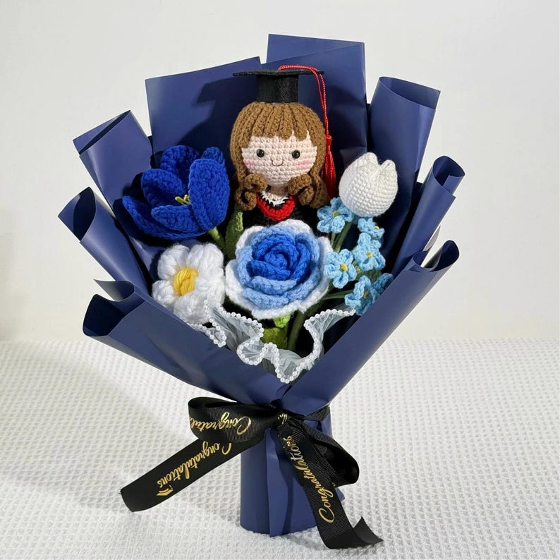 May include: A handcrafted bouquet featuring a crocheted graduate doll wearing a black cap and gown, surrounded by blue, white, and light blue crocheted flowers. The bouquet is wrapped in navy blue paper and tied with a black ribbon that says "Congratulations".