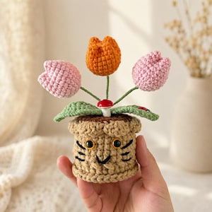May include: A crocheted tan pot with a cat-like face featuring black embroidered whiskers and eyes, holding three yarn flowers in orange and pink. A small red and white mushroom and a tiny ladybug are attached to the green crocheted leaves.