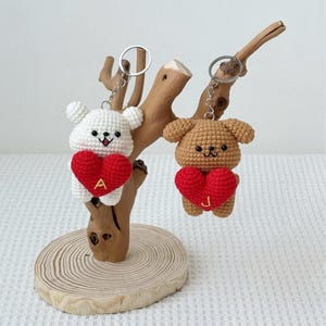 May include: Two crochet bear keychains, one white and one brown, each holding a red heart. The white bear has the letter "A" on its heart, and the brown bear has the letter "J". Both keychains are attached to silver keyrings.