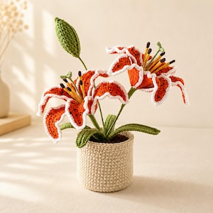 Puede incluir: Una planta de lirio tigre tejida a ganchillo con dos flores de color naranja vibrante con manchas marrón oscuro y bordes blancos, un capullo verde y hojas verdes, en una maceta de ganchillo beige con un patrón de puntos visible.