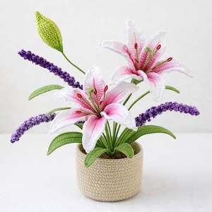 Pode incluir: Um arranjo floral de crochê com lírios rosa e brancos, lavanda roxa e um botão verde. As flores estão em um vaso de crochê bege. O trabalho artesanal mostra pontos detalhados e cores vibrantes, perfeito para decoração de casa.