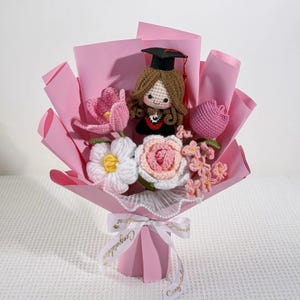 May include: A handmade crochet bouquet featuring a graduate doll in a black cap and gown, surrounded by pink, white, and peach-colored crochet flowers. The bouquet is wrapped in pink paper and tied with a white ribbon that says "Congratulations".