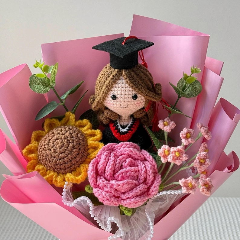 May include: A crocheted graduation bouquet featuring a doll wearing a black cap and gown. The bouquet includes a sunflower, a pink rose, and small pink flowers, all wrapped in pink paper.